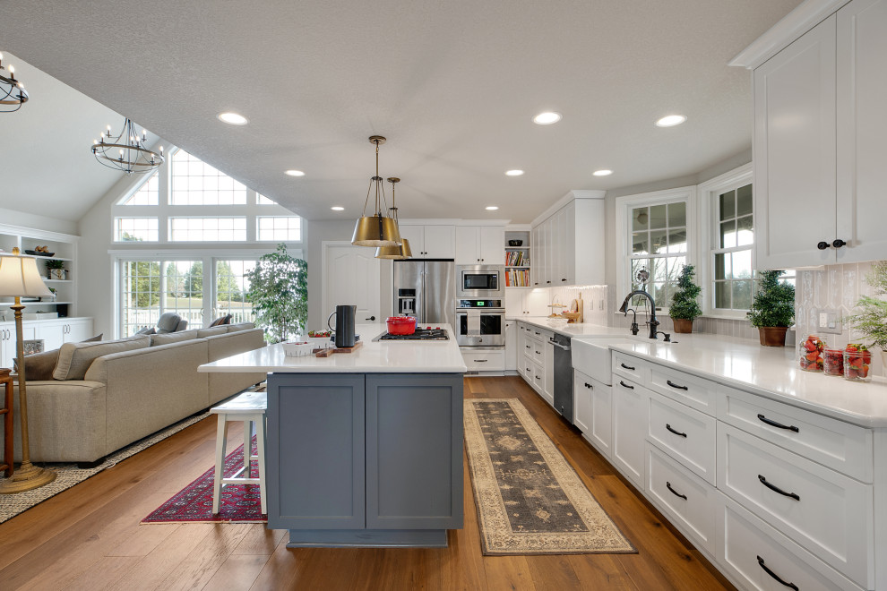 Design ideas for a large country u-shaped eat-in kitchen in Portland with a farmhouse sink, shaker cabinets, white cabinets, quartz benchtops, grey splashback, ceramic splashback, stainless steel appliances, medium hardwood floors, with island, brown floor and white benchtop.