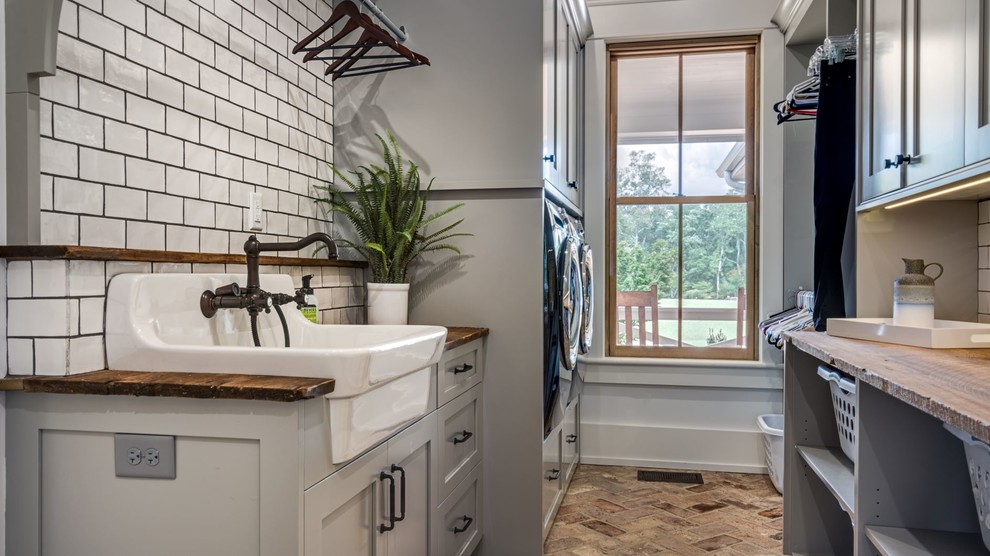 Photo of a mid-sized country galley dedicated laundry room in Nashville with a farmhouse sink, shaker cabinets, grey cabinets, wood benchtops, grey walls, brick floors, a side-by-side washer and dryer, brown floor and brown benchtop.