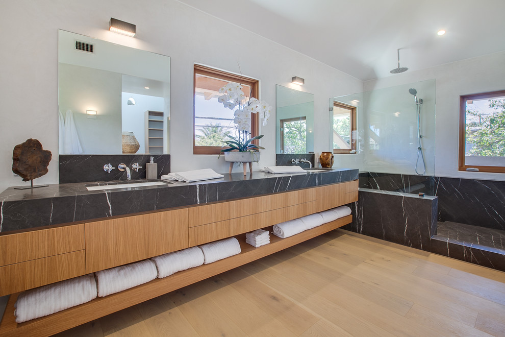 Tropical master bathroom in Los Angeles with flat-panel cabinets, medium wood cabinets, an open shower, white walls, light hardwood floors, an undermount sink, beige floor, an open shower and black benchtops.