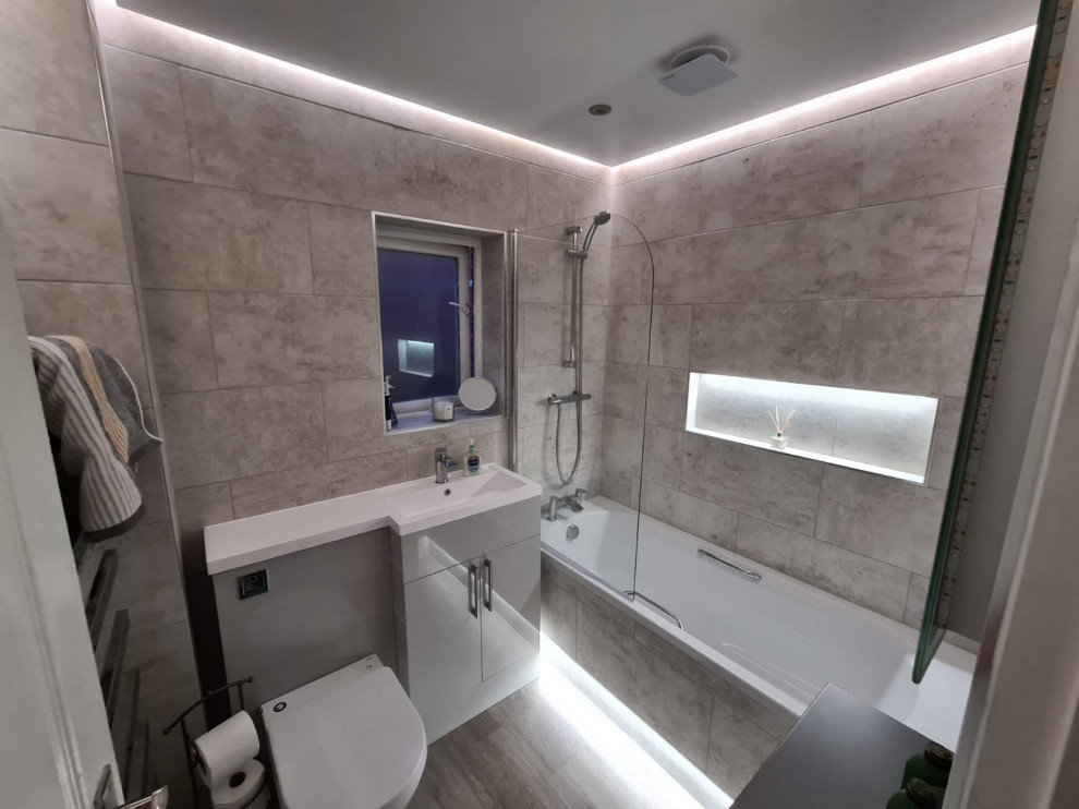 Photo of a mid-sized contemporary kids bathroom in Cheshire with flat-panel cabinets, grey cabinets, a drop-in tub, a shower/bathtub combo, a two-piece toilet, beige tile, stone tile, beige walls, laminate floors, a drop-in sink, solid surface benchtops, beige floor, a hinged shower door, white benchtops, a single vanity and a built-in vanity.