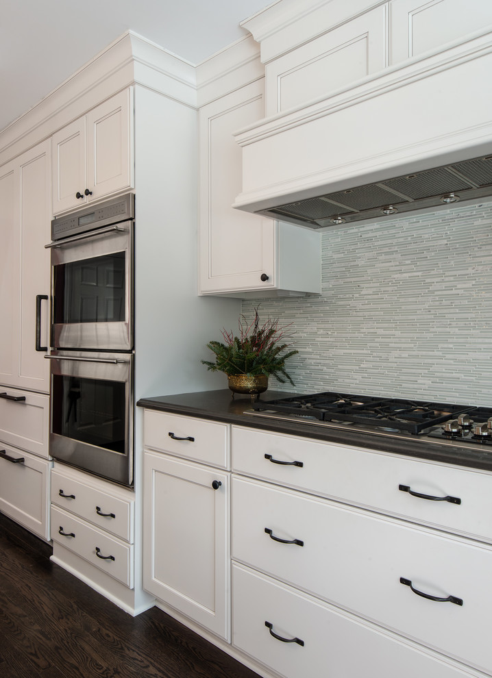 This is an example of a mid-sized transitional l-shaped open plan kitchen in Detroit with a farmhouse sink, recessed-panel cabinets, white cabinets, quartz benchtops, white splashback, glass tile splashback, stainless steel appliances, dark hardwood floors, with island, black floor and multi-coloured benchtop.