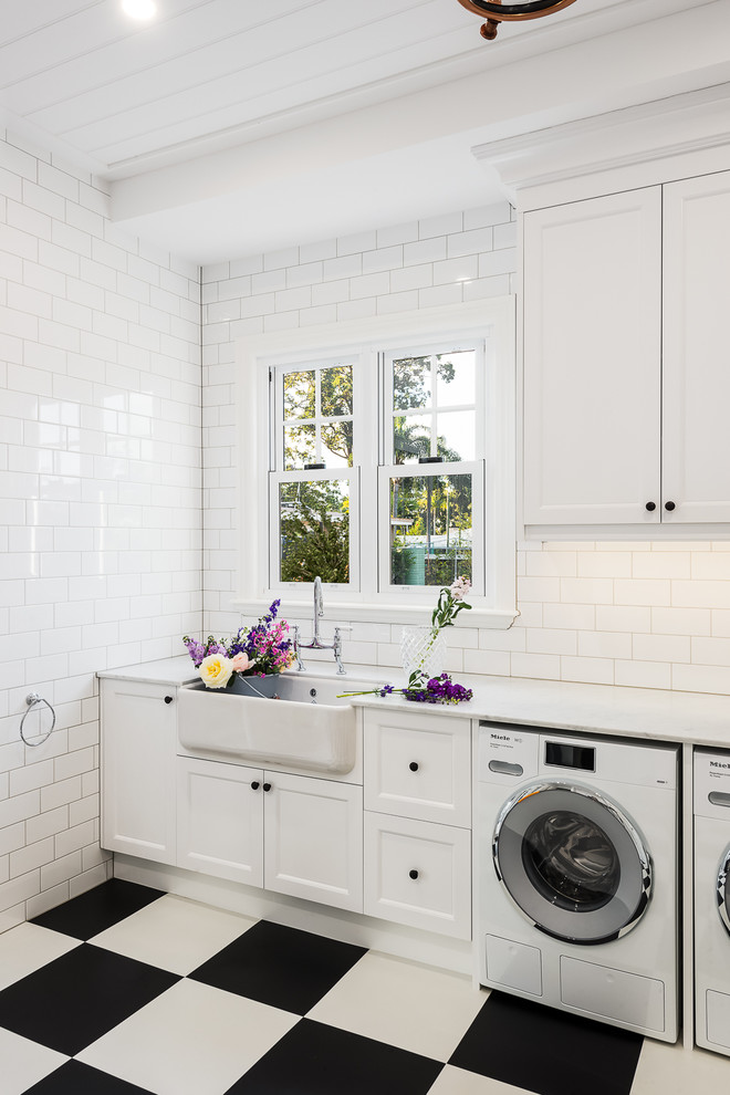 This is an example of a beach style single-wall dedicated laundry room in Gold Coast - Tweed with recessed-panel cabinets, white cabinets, white benchtop, a farmhouse sink, white walls, a side-by-side washer and dryer and multi-coloured floor.