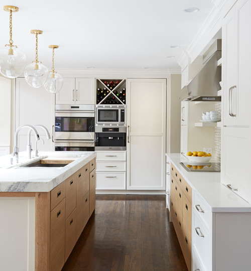 Bright galley kitchen with two-tone RTA cabinets—white uppers and warm wood drawer bases—marble countertops, globe brass pendants, and built-in wall ovens with wine cubby.