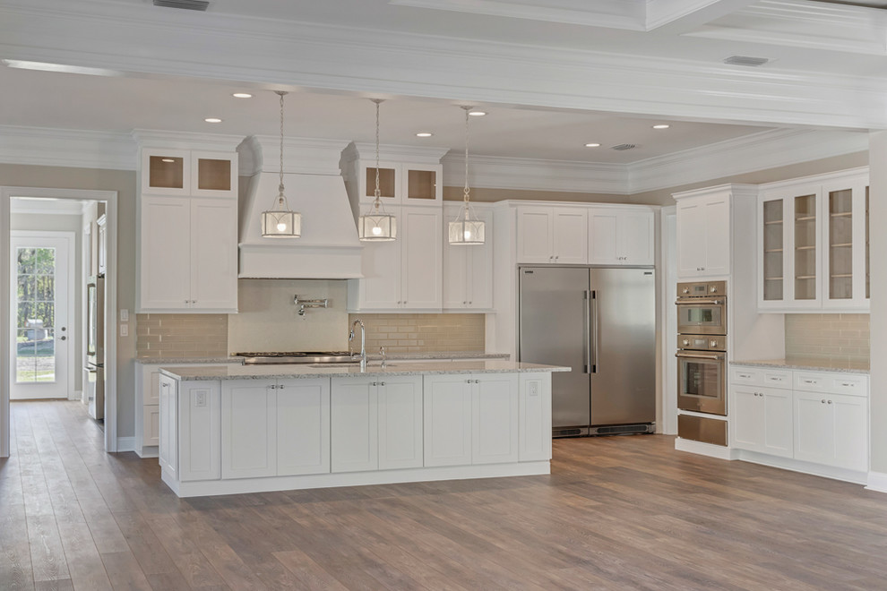 Inspiration for a large country single-wall eat-in kitchen in Jacksonville with a double-bowl sink, shaker cabinets, white cabinets, granite benchtops, beige splashback, glass tile splashback, stainless steel appliances, vinyl floors, with island, brown floor and beige benchtop.