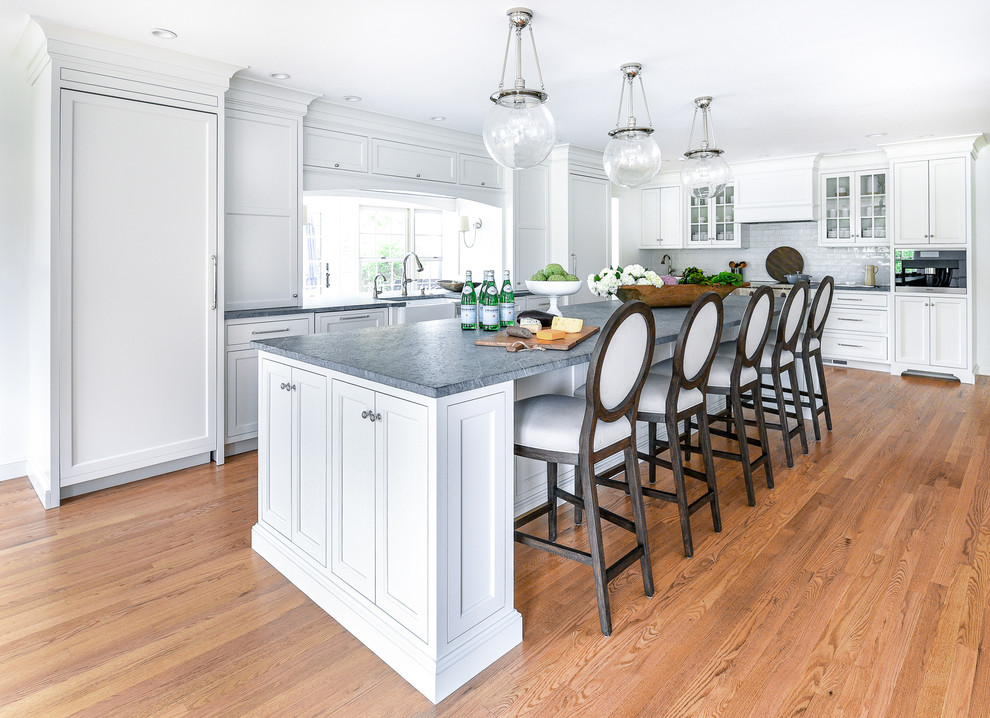 Design ideas for a large transitional l-shaped eat-in kitchen in Philadelphia with a farmhouse sink, flat-panel cabinets, white cabinets, soapstone benchtops, white splashback, ceramic splashback, panelled appliances, light hardwood floors, with island and grey benchtop.