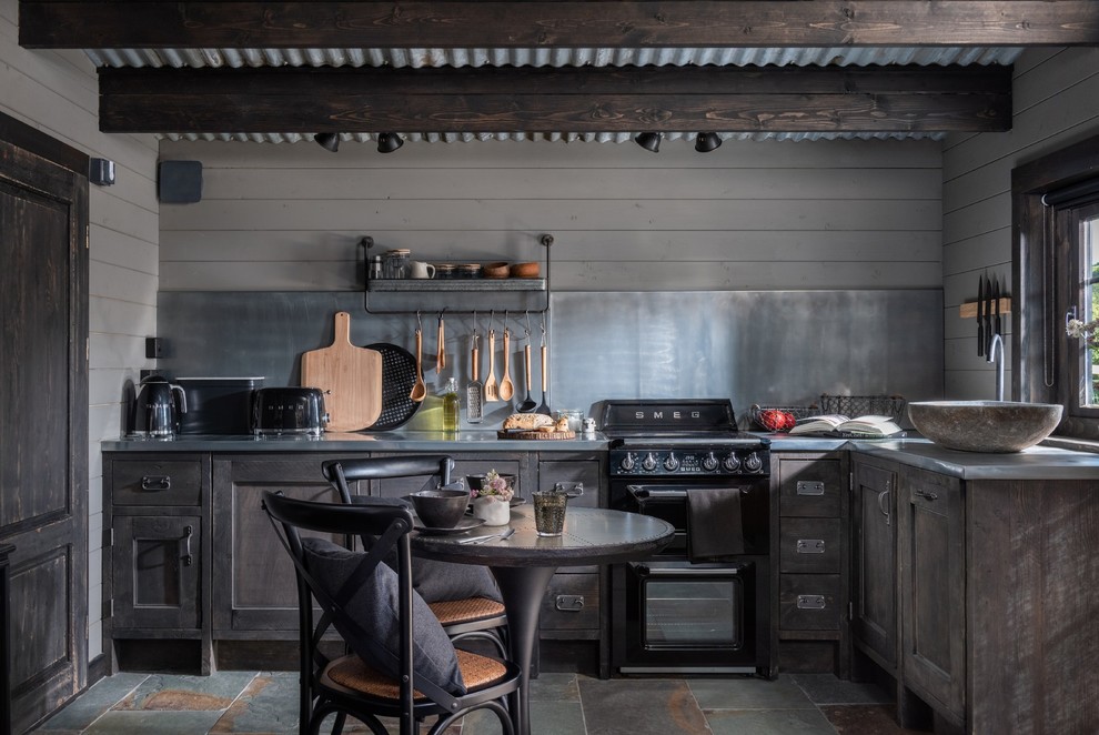 This is an example of a mid-sized country l-shaped eat-in kitchen in Cornwall with recessed-panel cabinets, dark wood cabinets, stainless steel benchtops, grey splashback, black appliances, slate floors, grey floor and grey benchtop.