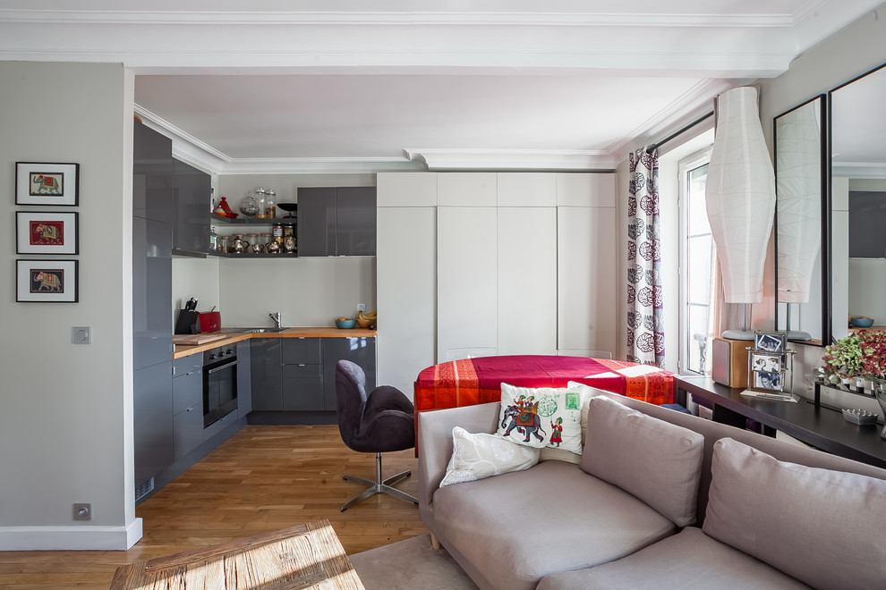 This is an example of a small contemporary l-shaped open plan kitchen in Paris with no island, beaded inset cabinets, grey cabinets, wood benchtops, grey splashback, glass sheet splashback, panelled appliances, a single-bowl sink and light hardwood floors.