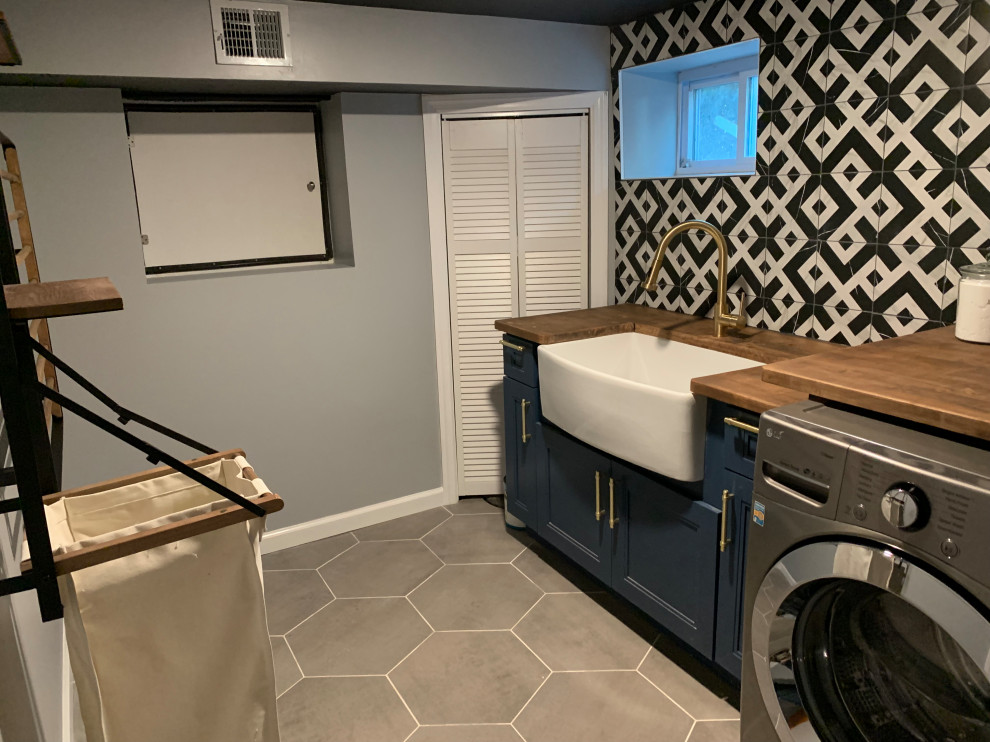 Photo of a mid-sized contemporary galley dedicated laundry room in New York with a farmhouse sink, beaded inset cabinets, blue cabinets, wood benchtops, multi-coloured splashback, cement tile splashback, grey walls, ceramic floors, a side-by-side washer and dryer, grey floor and brown benchtop.