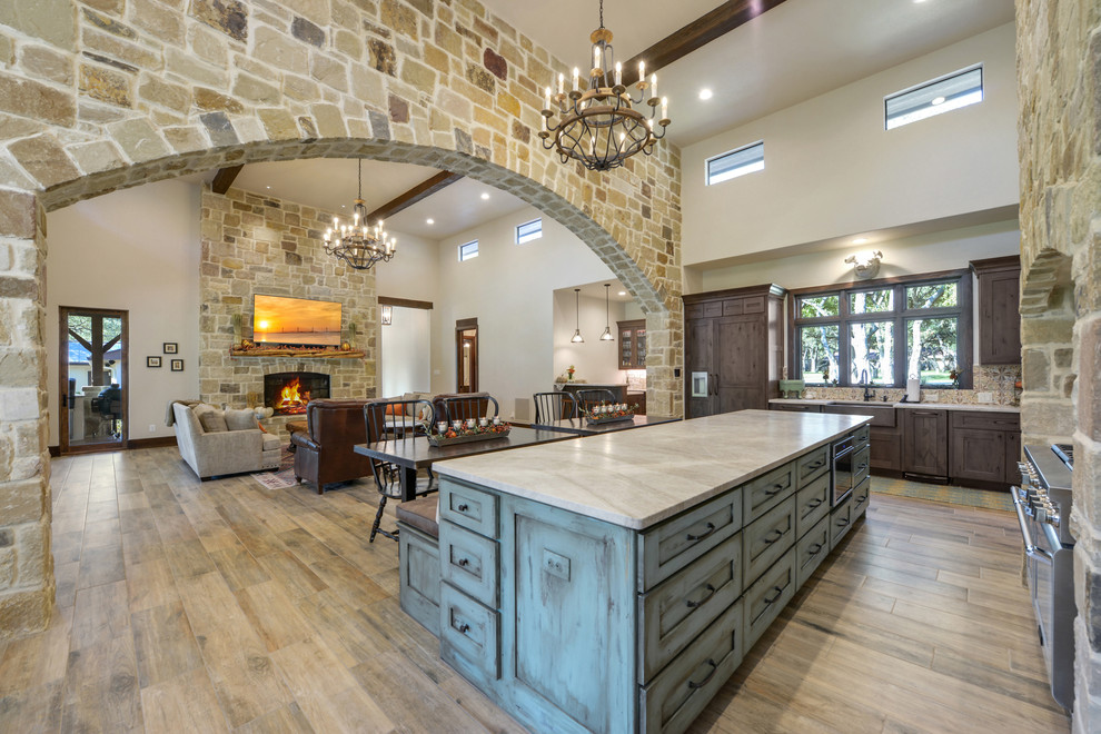 This is an example of a country l-shaped open plan kitchen in Austin with a farmhouse sink, shaker cabinets, distressed cabinets, granite benchtops, multi-coloured splashback, porcelain splashback, panelled appliances, ceramic floors, with island, brown floor and beige benchtop.