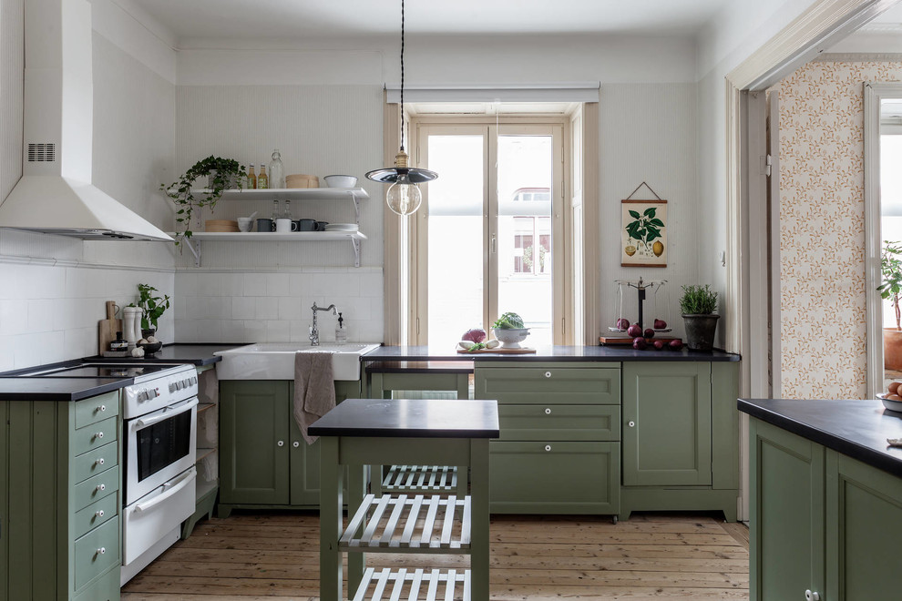 Photo of a mid-sized country separate kitchen in Stockholm with a farmhouse sink, shaker cabinets, green cabinets, white splashback, white appliances, light hardwood floors, with island, beige floor and black benchtop.