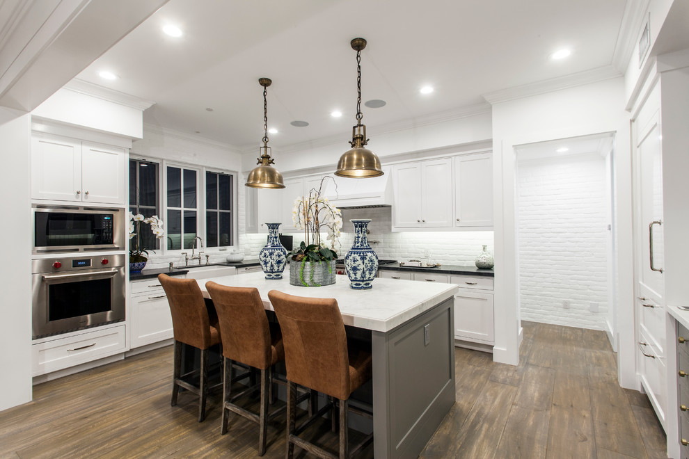 Photo of a mid-sized beach style l-shaped eat-in kitchen in Orange County with shaker cabinets, white cabinets, marble benchtops, white splashback, stainless steel appliances, dark hardwood floors and with island.