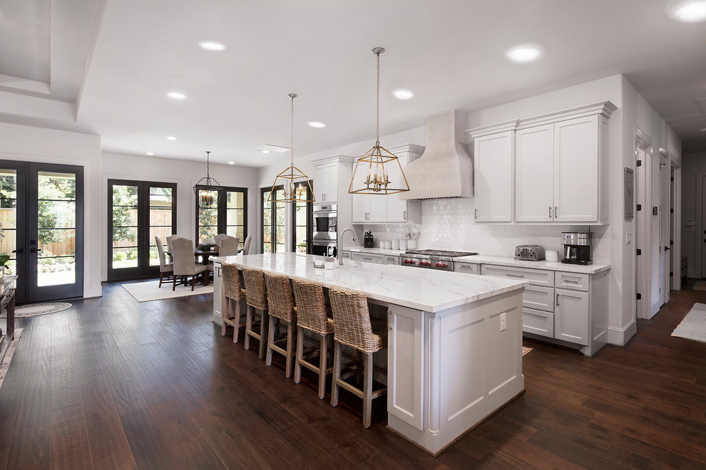 Photo of an expansive traditional single-wall open plan kitchen in Houston with a farmhouse sink, white cabinets, marble benchtops, white splashback, subway tile splashback, stainless steel appliances, dark hardwood floors, with island, brown floor and white benchtop.