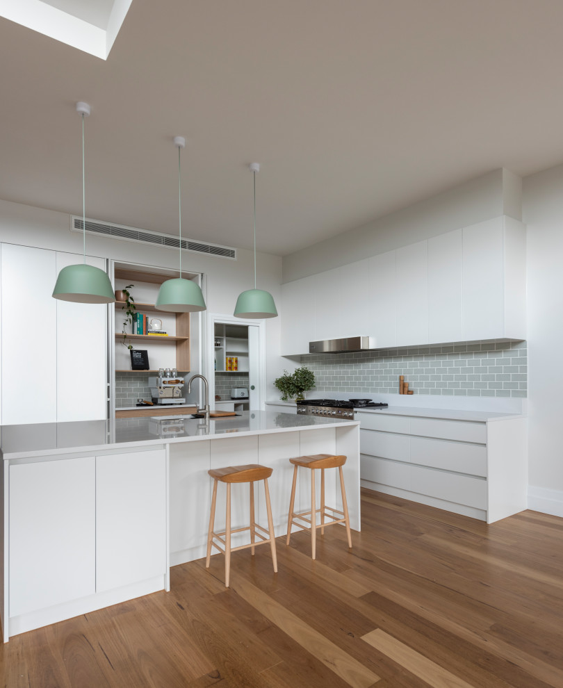 Inspiration for a large contemporary l-shaped kitchen in Melbourne with a double-bowl sink, flat-panel cabinets, white cabinets, quartz benchtops, green splashback, subway tile splashback, stainless steel appliances, medium hardwood floors, with island, brown floor and white benchtop.
