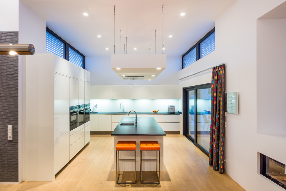 This is an example of a large contemporary single-wall open plan kitchen in Frankfurt with flat-panel cabinets, white cabinets, granite benchtops, white splashback, black appliances, medium hardwood floors, with island and a single-bowl sink.