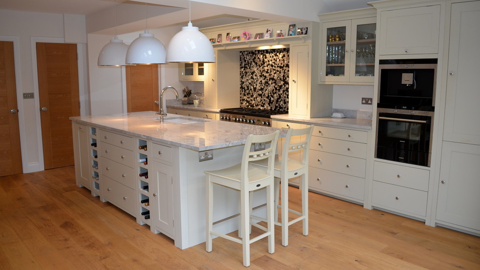 Inspiration for a mid-sized contemporary single-wall eat-in kitchen in Hertfordshire with a farmhouse sink, beaded inset cabinets, white cabinets, solid surface benchtops, black splashback, ceramic splashback, black appliances, painted wood floors and with island.