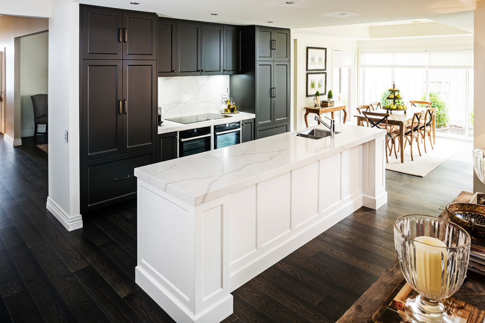 This is an example of a large transitional galley open plan kitchen in Melbourne with a double-bowl sink, recessed-panel cabinets, brown cabinets, quartz benchtops, white splashback, stone slab splashback, black appliances, dark hardwood floors, with island, brown floor and white benchtop.