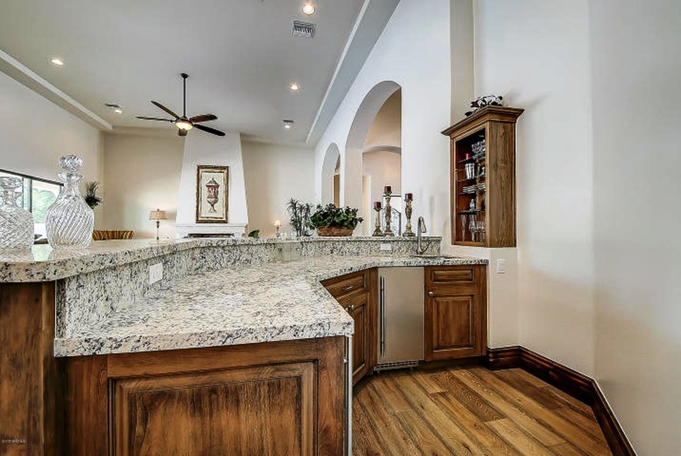 Inspiration for a mid-sized mediterranean u-shaped seated home bar in Phoenix with an undermount sink, raised-panel cabinets, medium wood cabinets, granite benchtops, white splashback, medium hardwood floors, brown floor and white benchtop.