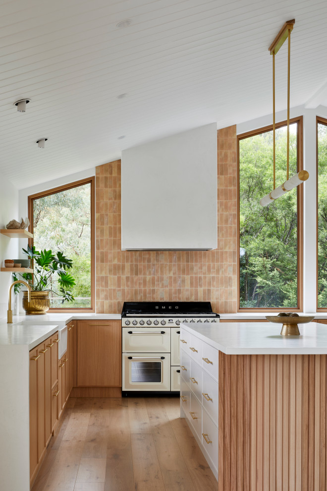 This is an example of a large modern u-shaped open plan kitchen in Melbourne with a farmhouse sink, shaker cabinets, medium wood cabinets, pink splashback, mosaic tile splashback, black appliances, medium hardwood floors, with island, brown floor, white benchtop and exposed beam.