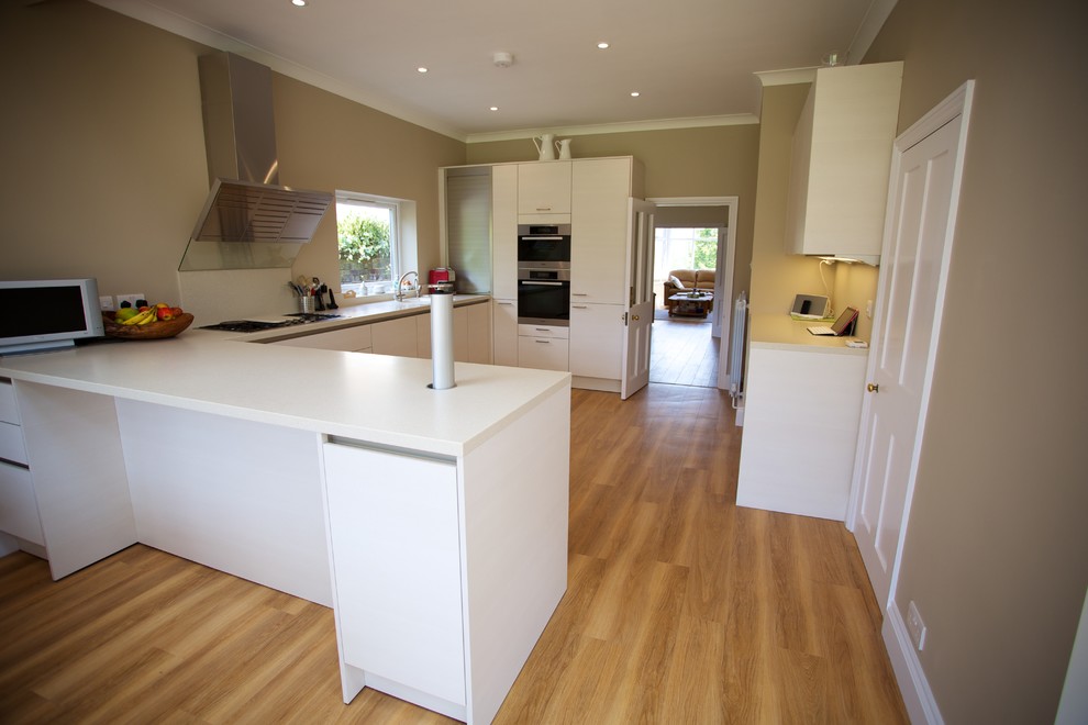 Mid-sized contemporary u-shaped eat-in kitchen in Hampshire with an integrated sink, flat-panel cabinets, white cabinets, solid surface benchtops, white splashback, stainless steel appliances, medium hardwood floors and a peninsula.