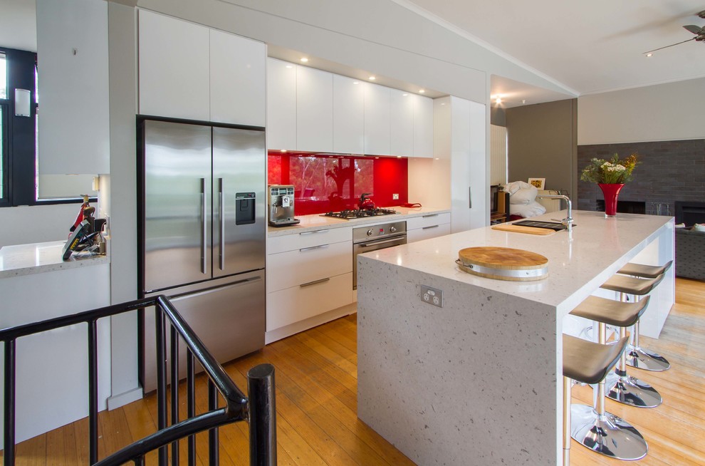 This is an example of a mid-sized contemporary galley open plan kitchen in Melbourne with flat-panel cabinets, white cabinets, quartz benchtops, red splashback, glass sheet splashback, stainless steel appliances, medium hardwood floors and with island.