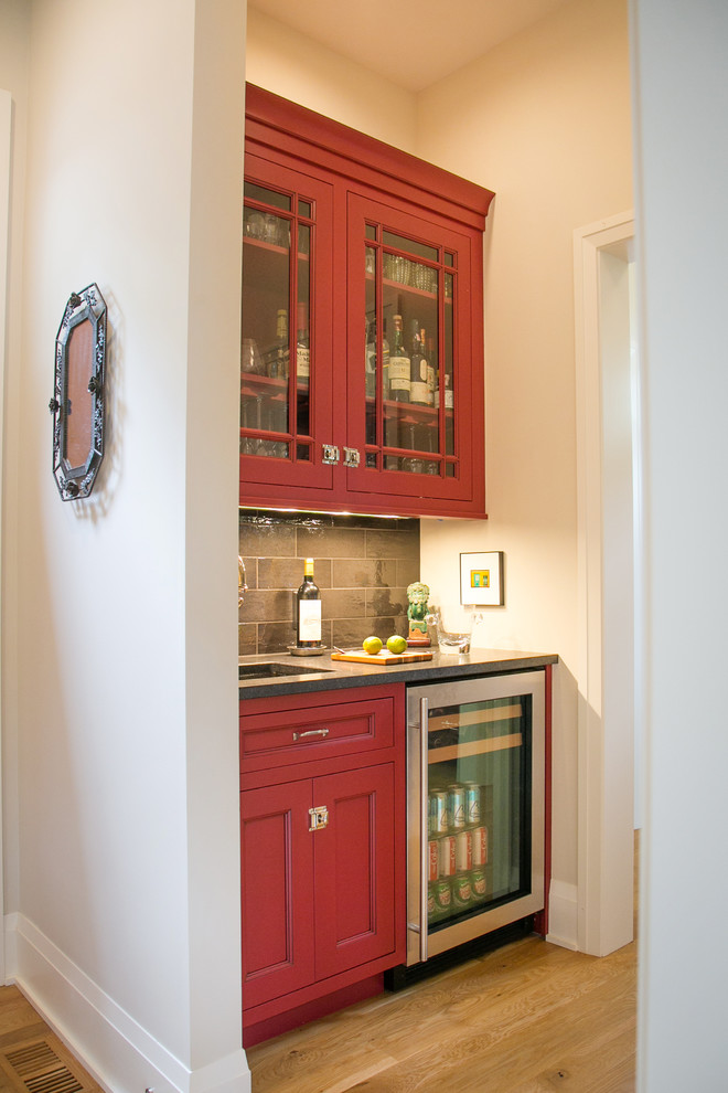 Inspiration for a small arts and crafts galley wet bar in Milwaukee with an undermount sink, recessed-panel cabinets, red cabinets, granite benchtops, black splashback, porcelain splashback, light hardwood floors, brown floor and black benchtop.