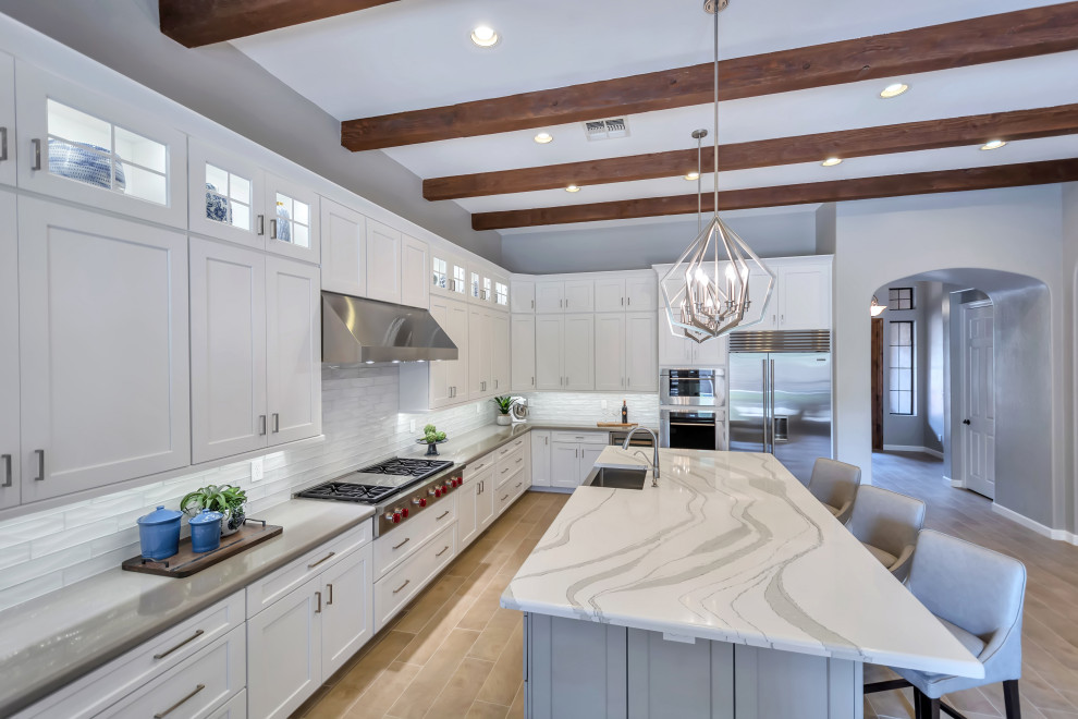 Large transitional l-shaped porcelain tile and brown floor eat-in kitchen photo in Phoenix with a farmhouse sink, shaker cabinets, white cabinets, quartz countertops, white backsplash, subway tile backsplash, stainless steel appliances, an island and gray countertops