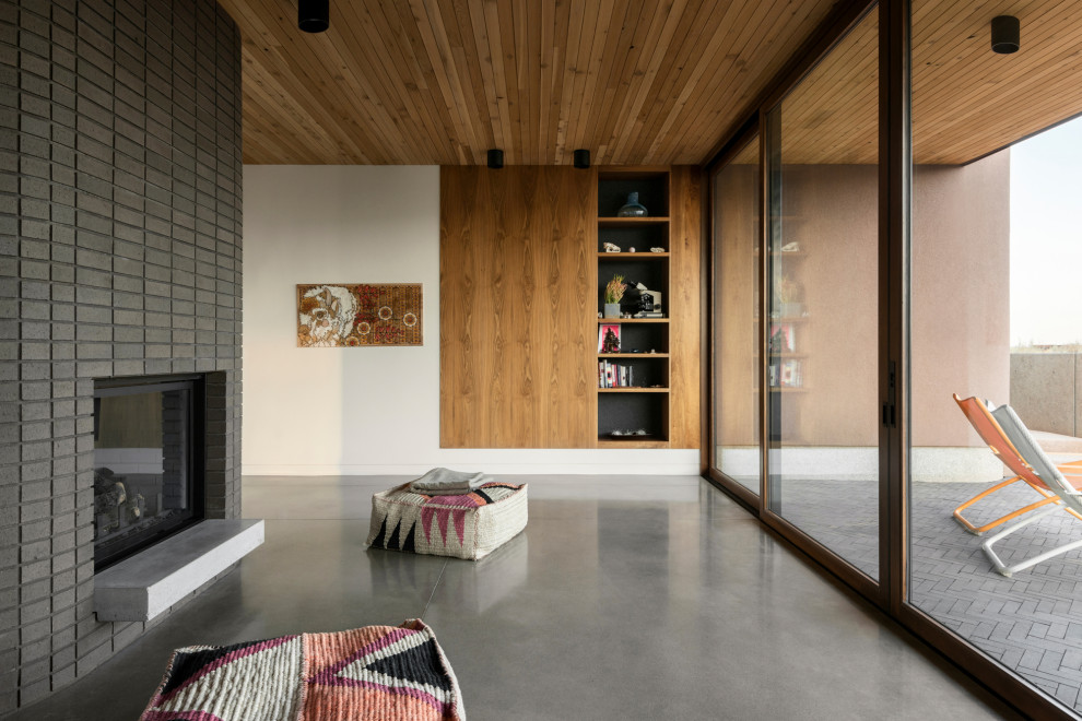 На фото: открытая гостиная комната с с книжными шкафами и полками, белыми стенами, бетонным полом, двусторонним камином, фасадом камина из кирпича, серым полом, деревянным потолком и деревянными стенами с