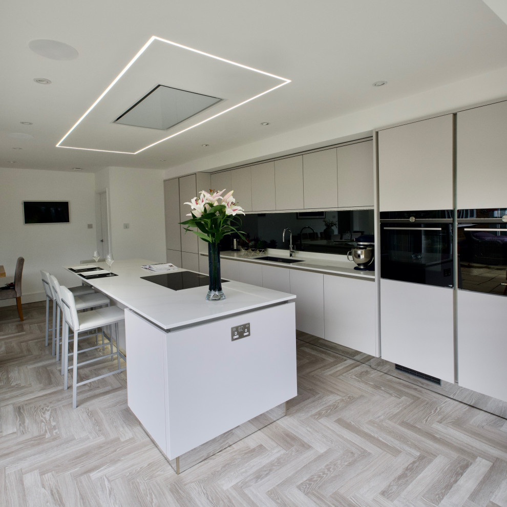На фото: кухня-гостиная: освещение в стиле модернизм с врезной мойкой, плоскими фасадами, серым фартуком, зеркальным фартуком, черной техникой, островом и белой столешницей с