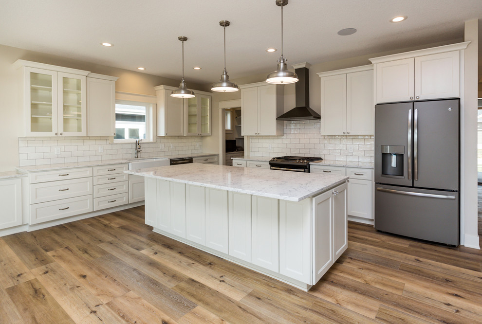 Inspiration for a country l-shaped eat-in kitchen in Other with a farmhouse sink, shaker cabinets, white cabinets, quartz benchtops, white splashback, ceramic splashback, stainless steel appliances, vinyl floors, with island and brown floor.