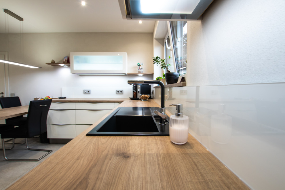 Photo of a mid-sized contemporary u-shaped separate kitchen in Dusseldorf with a drop-in sink, flat-panel cabinets, white cabinets, wood benchtops, white splashback, glass sheet splashback, stainless steel appliances, cement tiles, no island, grey floor and brown benchtop.