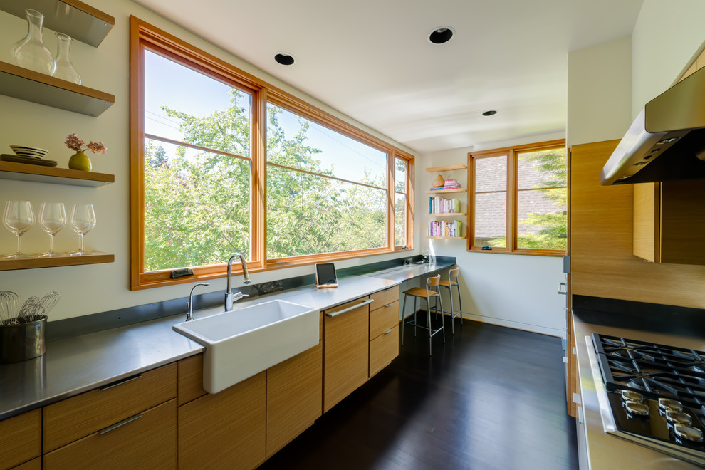 This is an example of a contemporary galley separate kitchen in Portland with a farmhouse sink, flat-panel cabinets, light wood cabinets, stainless steel benchtops, grey splashback, stainless steel appliances, dark hardwood floors and no island.