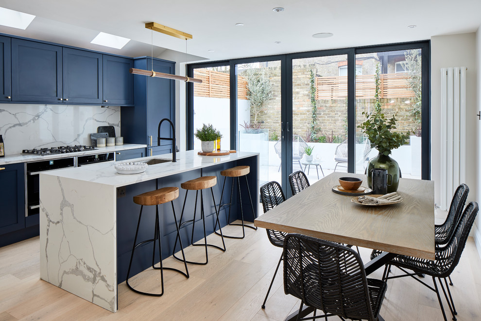 This is an example of a contemporary galley eat-in kitchen in London with an undermount sink, shaker cabinets, blue cabinets, white splashback, stone slab splashback, panelled appliances, light hardwood floors, with island, beige floor and white benchtop.