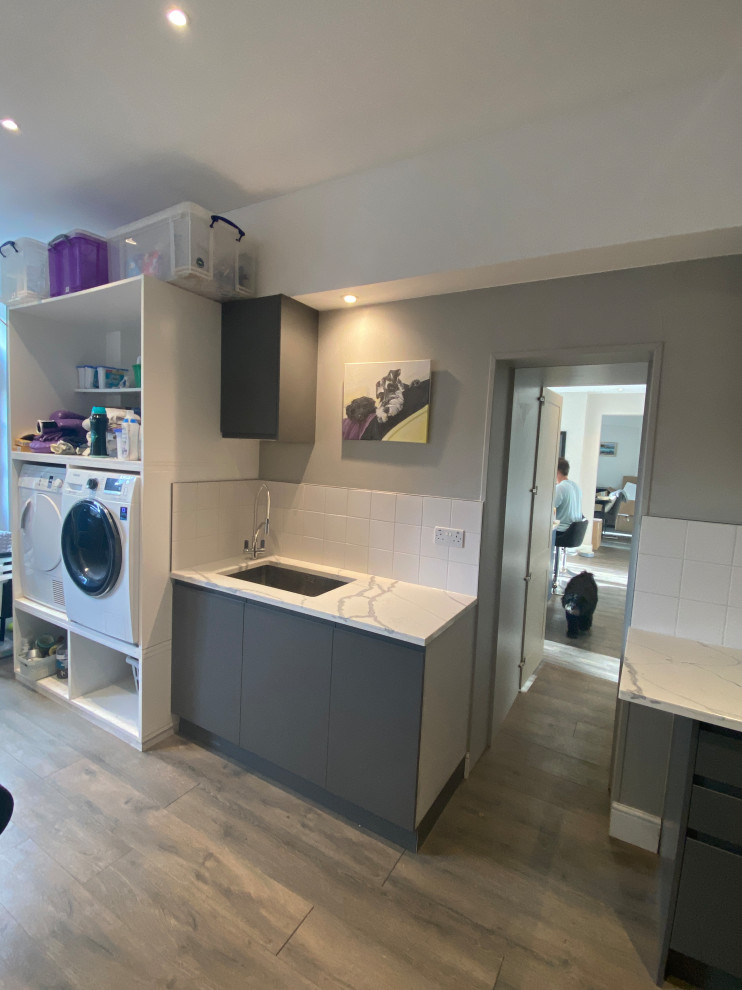 ロンドンにある高級な広いコンテンポラリースタイルのおしゃれな家事室 (ll型、ドロップインシンク、フラットパネル扉のキャビネット、グレーのキャビネット、珪岩カウンター、白いキッチンパネル、磁器タイルのキッチンパネル、ベージュの壁、ラミネートの床、左右配置の洗濯機・乾燥機、グレーの床、マルチカラーのキッチンカウンター) の写真