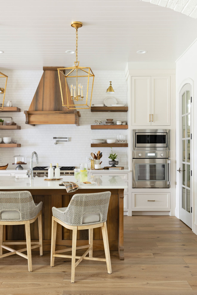 Inredning av ett modernt stort vit vitt kök, med en nedsänkt diskho, luckor med profilerade fronter, vita skåp, granitbänkskiva, vitt stänkskydd, stänkskydd i stenkakel, rostfria vitvaror, mellanmörkt trägolv, en köksö och brunt golv