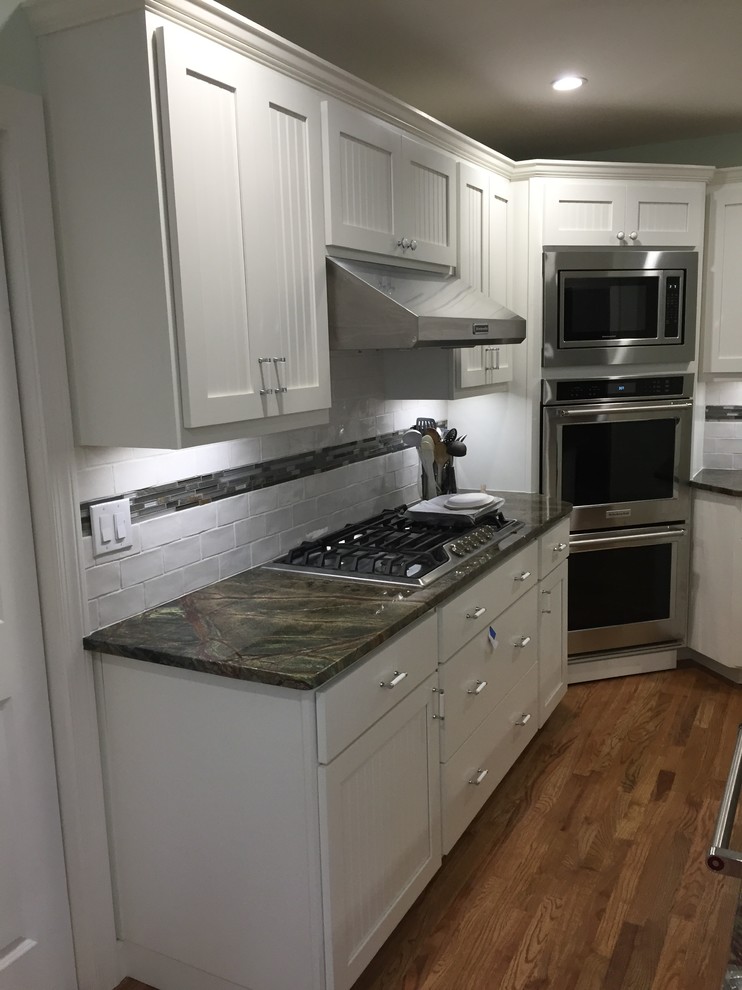 Example of a mid-sized transitional l-shaped medium tone wood floor and brown floor kitchen design in Atlanta with a farmhouse sink, shaker cabinets, white cabinets, marble countertops, white backsplash, ceramic backsplash, stainless steel appliances and an island