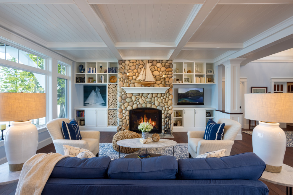 Cette photo montre un grand salon bord de mer ouvert avec un mur bleu, parquet foncé, une cheminée standard, un manteau de cheminée en pierre, un téléviseur encastré, un sol marron et un plafond à caissons.