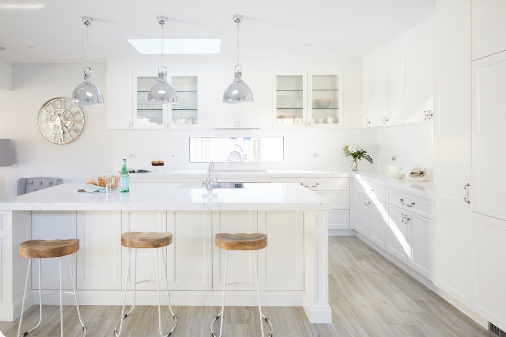 Photo of a transitional l-shaped kitchen in Sydney with an undermount sink, shaker cabinets, white cabinets, white splashback, panelled appliances, light hardwood floors, with island, beige floor and white benchtop.