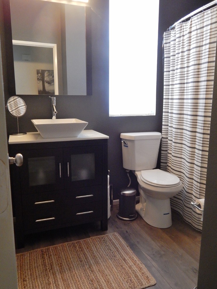 This is an example of a mid-sized contemporary bathroom in Los Angeles with glass-front cabinets, brown cabinets, an alcove tub, a shower/bathtub combo, a two-piece toilet, grey walls, laminate floors, a vessel sink, marble benchtops, grey floor, a shower curtain and white benchtops.