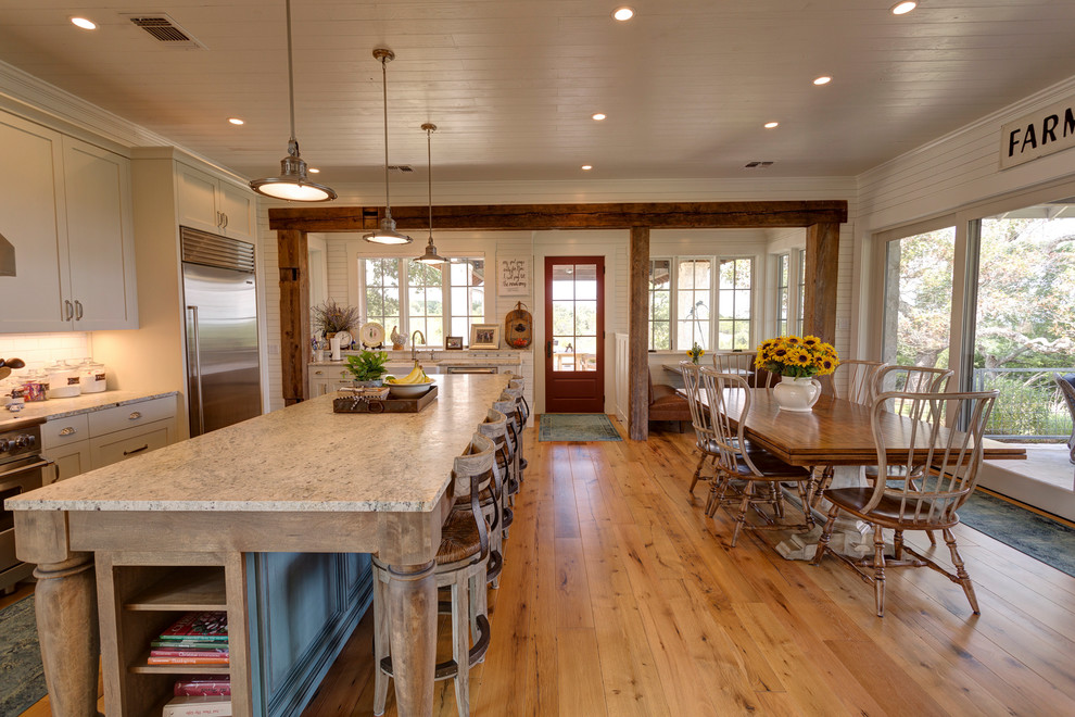 This is an example of a large country l-shaped eat-in kitchen in Austin with a farmhouse sink, shaker cabinets, white cabinets, granite benchtops, white splashback, ceramic splashback, stainless steel appliances, medium hardwood floors, with island and brown floor.