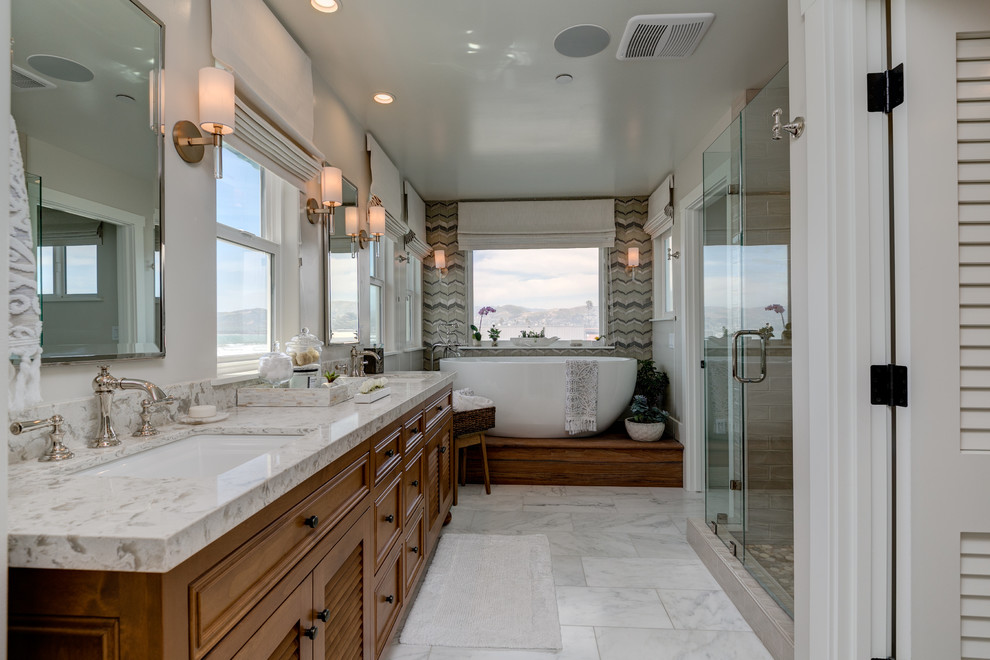 Photo of a mid-sized beach style master bathroom in Los Angeles with recessed-panel cabinets, medium wood cabinets, a freestanding tub, a one-piece toilet, gray tile, glass sheet wall, grey walls, marble floors, an undermount sink, quartzite benchtops, white floor and a shower curtain.