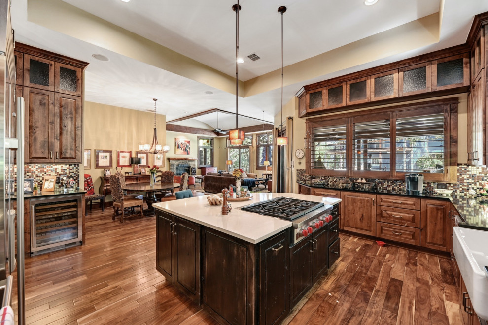 Photo of a mid-sized transitional u-shaped eat-in kitchen in Austin with a farmhouse sink, raised-panel cabinets, dark wood cabinets, quartzite benchtops, multi-coloured splashback, ceramic splashback, stainless steel appliances, medium hardwood floors, with island, brown floor, black benchtop and recessed.