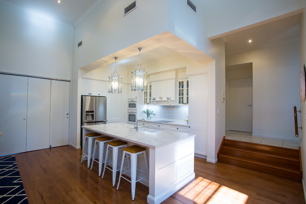 Photo of a large traditional galley kitchen pantry in Brisbane with a farmhouse sink, shaker cabinets, white cabinets, quartz benchtops, white splashback, subway tile splashback, white appliances, medium hardwood floors, with island, brown floor and white benchtop.