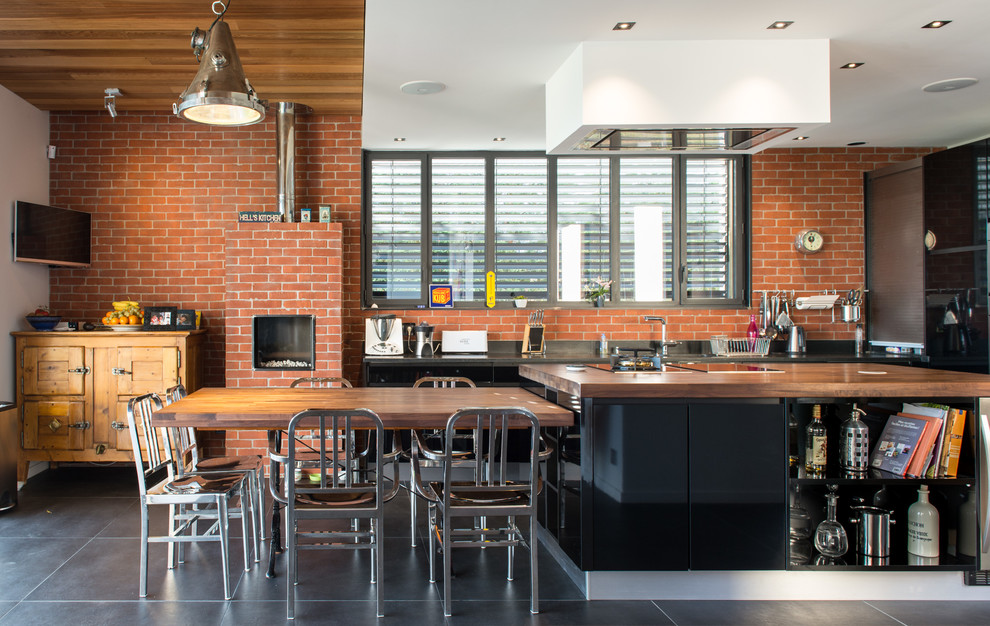 This is an example of a large industrial galley eat-in kitchen in Lyon with flat-panel cabinets, dark wood cabinets, wood benchtops, red splashback, black appliances, concrete floors and with island.