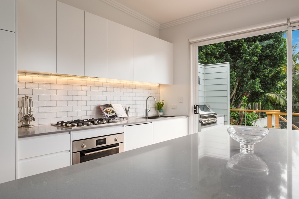 Photo of a mid-sized modern galley eat-in kitchen in Sydney with an integrated sink, white cabinets, white splashback, ceramic splashback, stainless steel appliances, with island, flat-panel cabinets, quartz benchtops and grey benchtop.