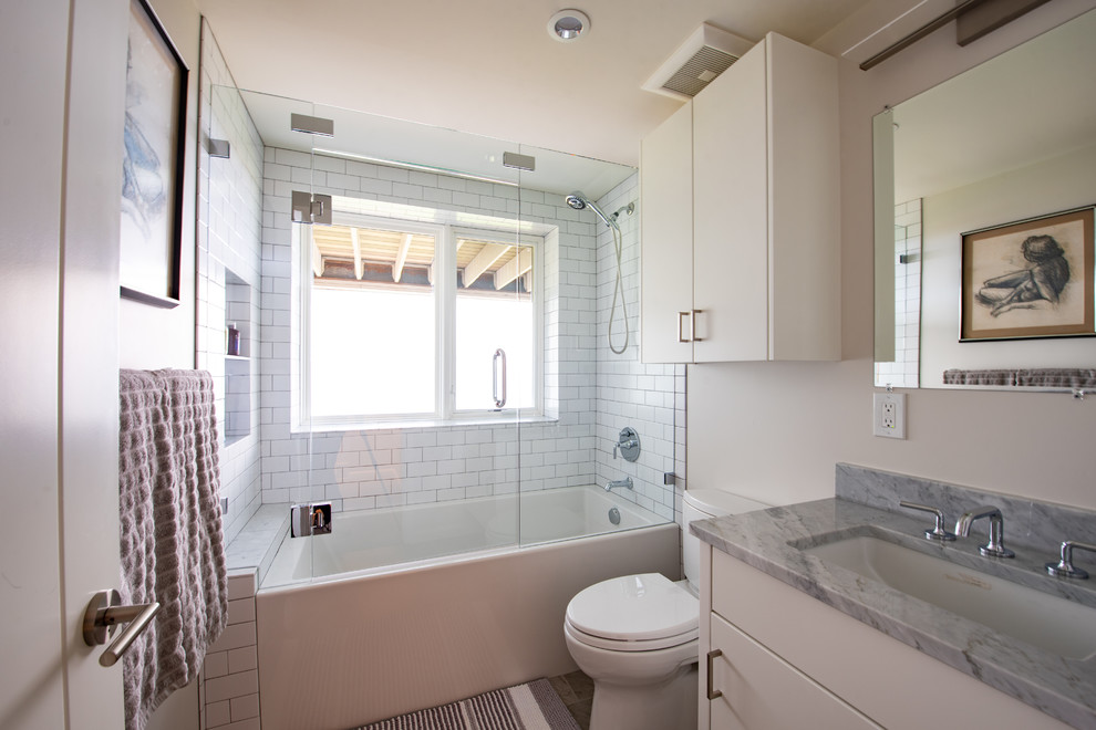 This is an example of a mid-sized midcentury 3/4 bathroom in Salt Lake City with flat-panel cabinets, white cabinets, an alcove tub, a shower/bathtub combo, a two-piece toilet, white tile, subway tile, white walls, an undermount sink, granite benchtops, a hinged shower door and grey benchtops.