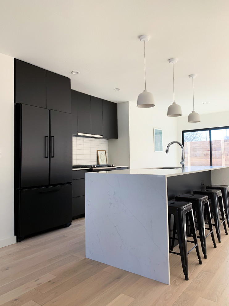 Contemporary galley kitchen in Austin with an undermount sink, flat-panel cabinets, black cabinets, white splashback, black appliances, light hardwood floors, with island, beige floor and white benchtop.
