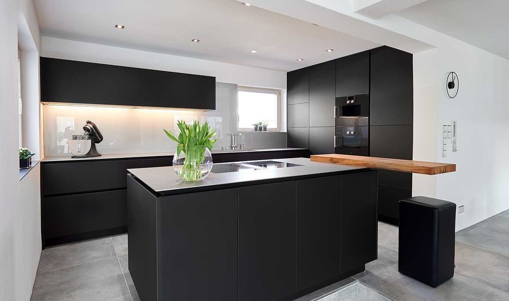 Design ideas for a contemporary l-shaped open plan kitchen in Stuttgart with a drop-in sink, flat-panel cabinets, black cabinets, stainless steel benchtops, white splashback, glass sheet splashback, black appliances, concrete floors, with island and grey floor.
