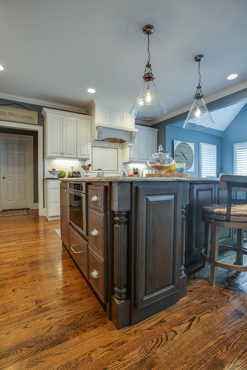 Large transitional l-shaped medium tone wood floor and brown floor open concept kitchen photo in Charlotte with white cabinets, granite countertops, white backsplash, stainless steel appliances, an island, a farmhouse sink, raised-panel cabinets, porcelain backsplash and white countertops