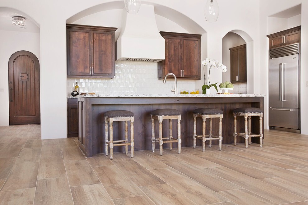 Open concept kitchen - mid-sized transitional l-shaped medium tone wood floor and brown floor open concept kitchen idea in Phoenix with an undermount sink, shaker cabinets, brown cabinets, marble countertops, white backsplash, porcelain backsplash, stainless steel appliances and an island
