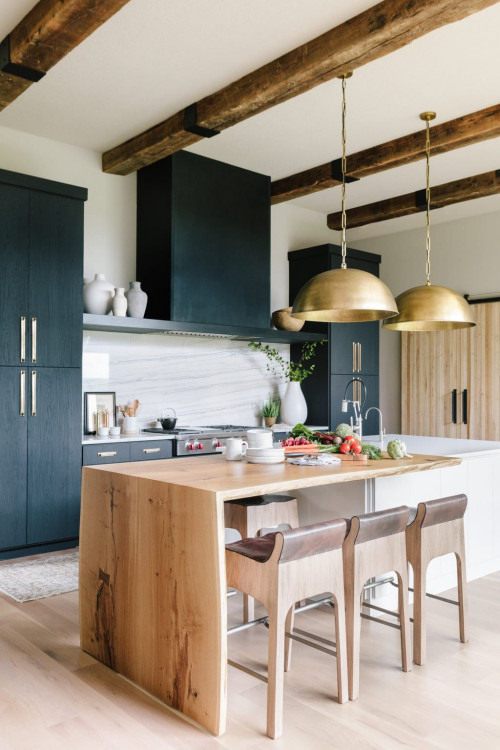 Modern open-concept kitchen with wood island, exposed beams, brass pendants and dark blue kitchen cabinets with tall pantry storage.